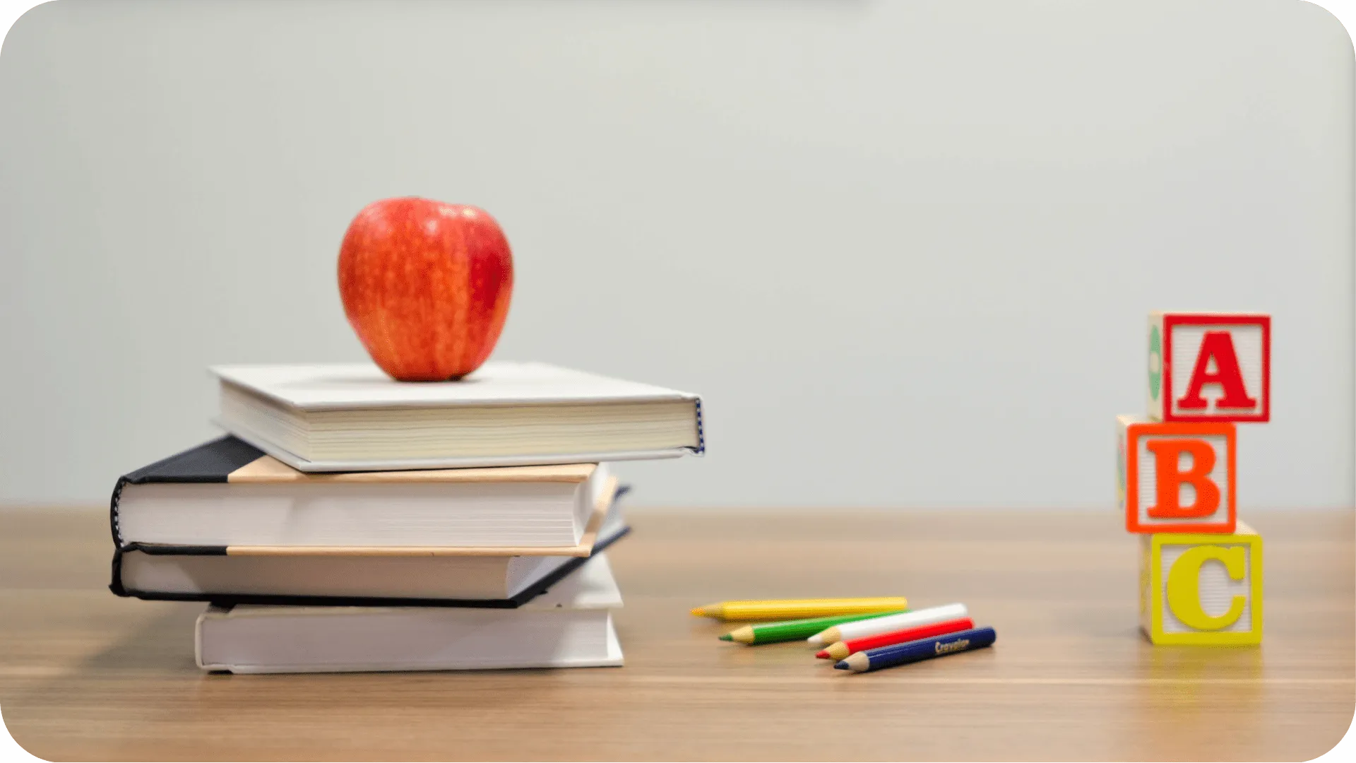 An apple on a pile of books, next to children's alphabet blocks and coloured pencils on a teachers desk in a school.