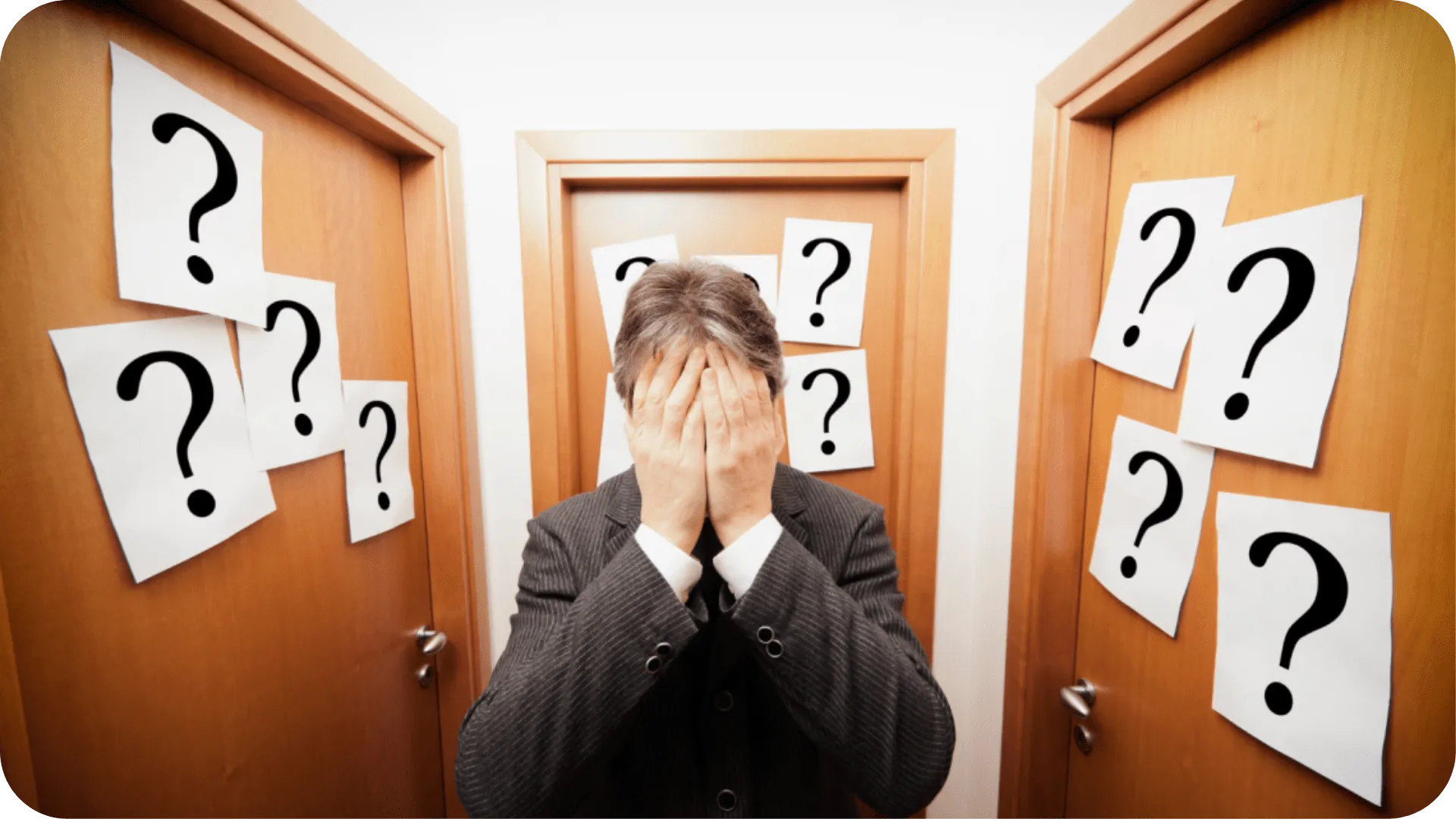 A man in a suit, looking confused with his hands on his face, in a hallway of closed doors with question parks on.
