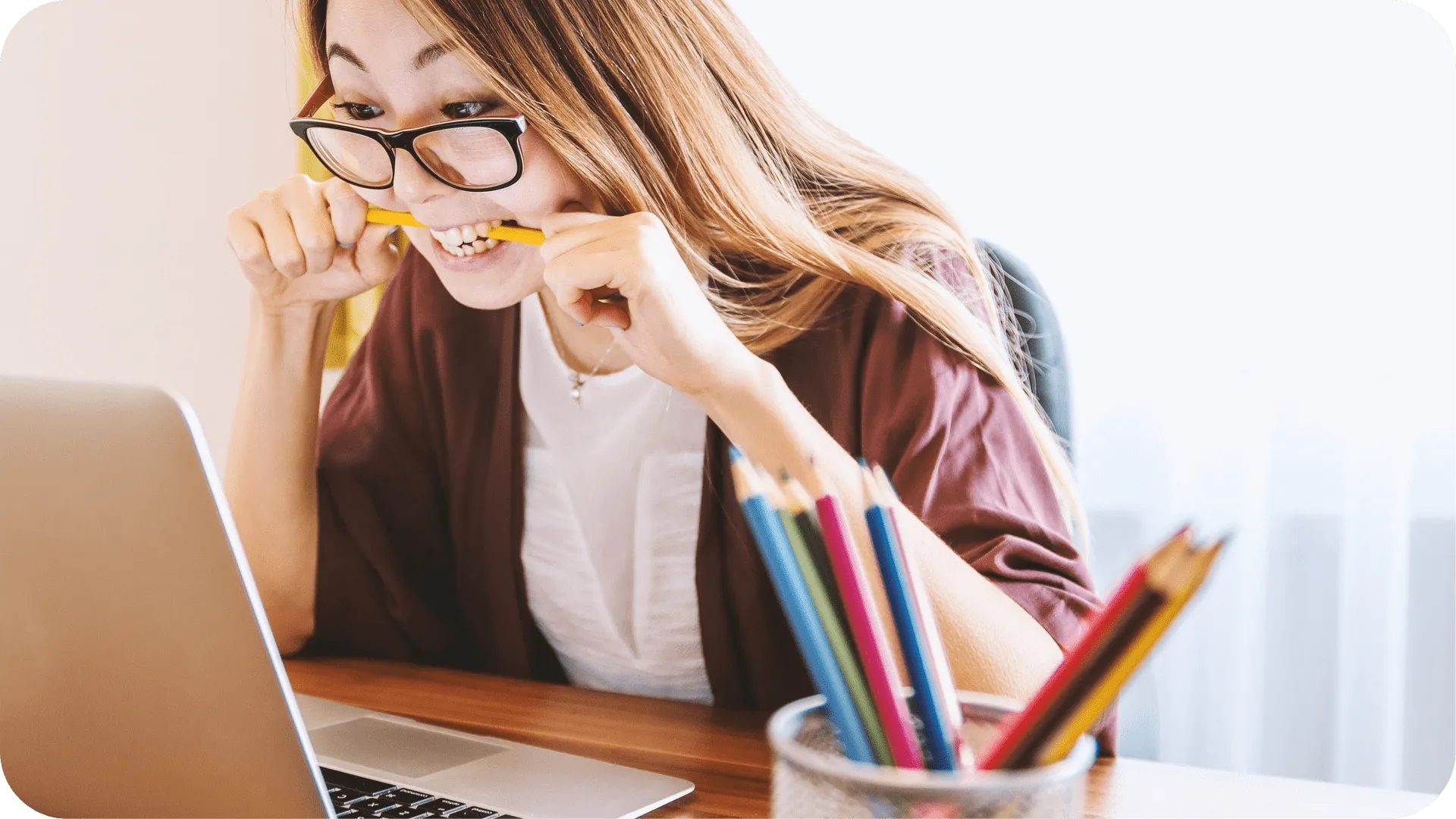Woman working on laptop holding a pencil in hands and in mouth.