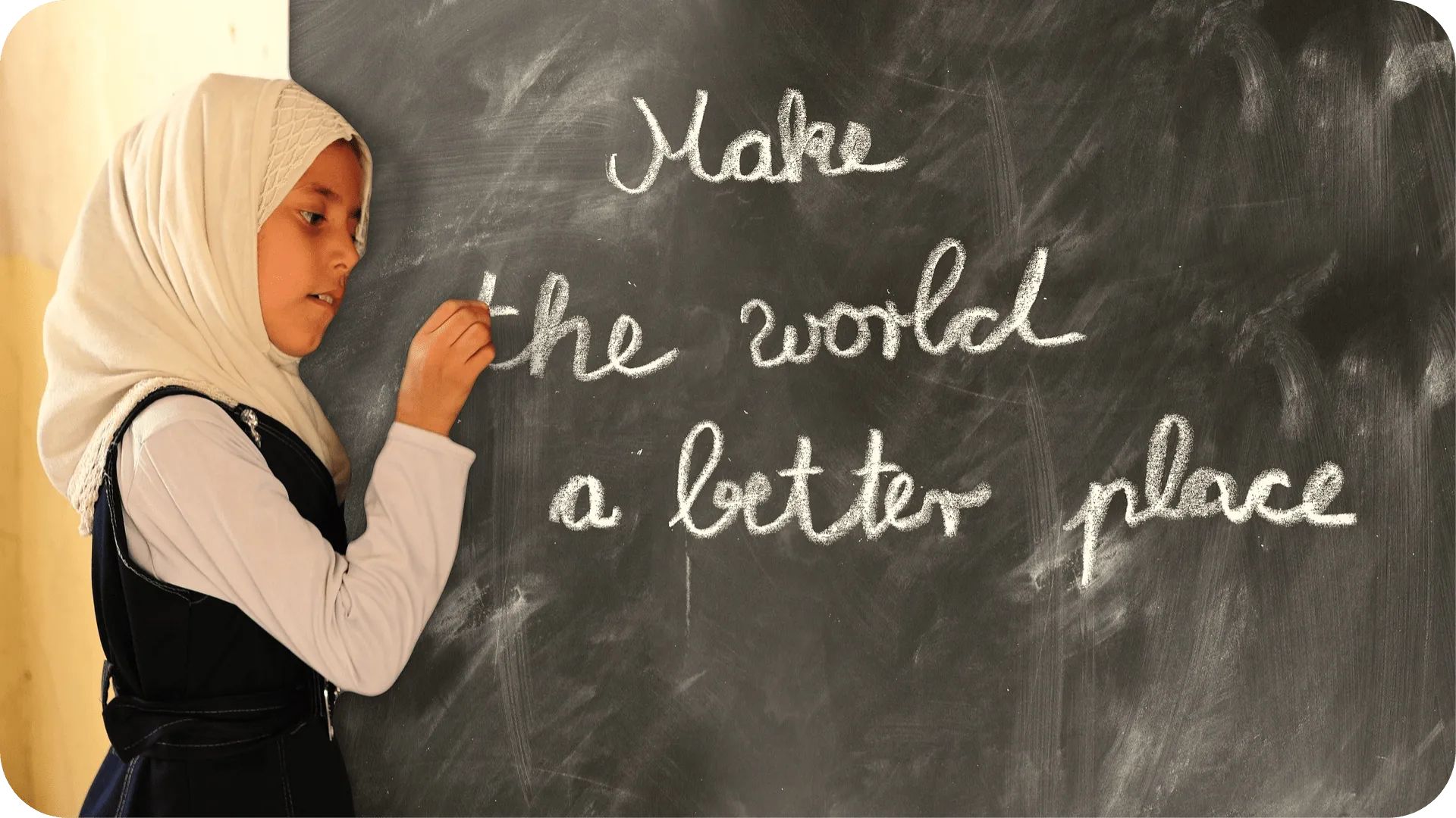 A young child in school draws on a chalkboard with the message 'The world is a better place with you in it,' expressing hope and a desire to make a positive impact.